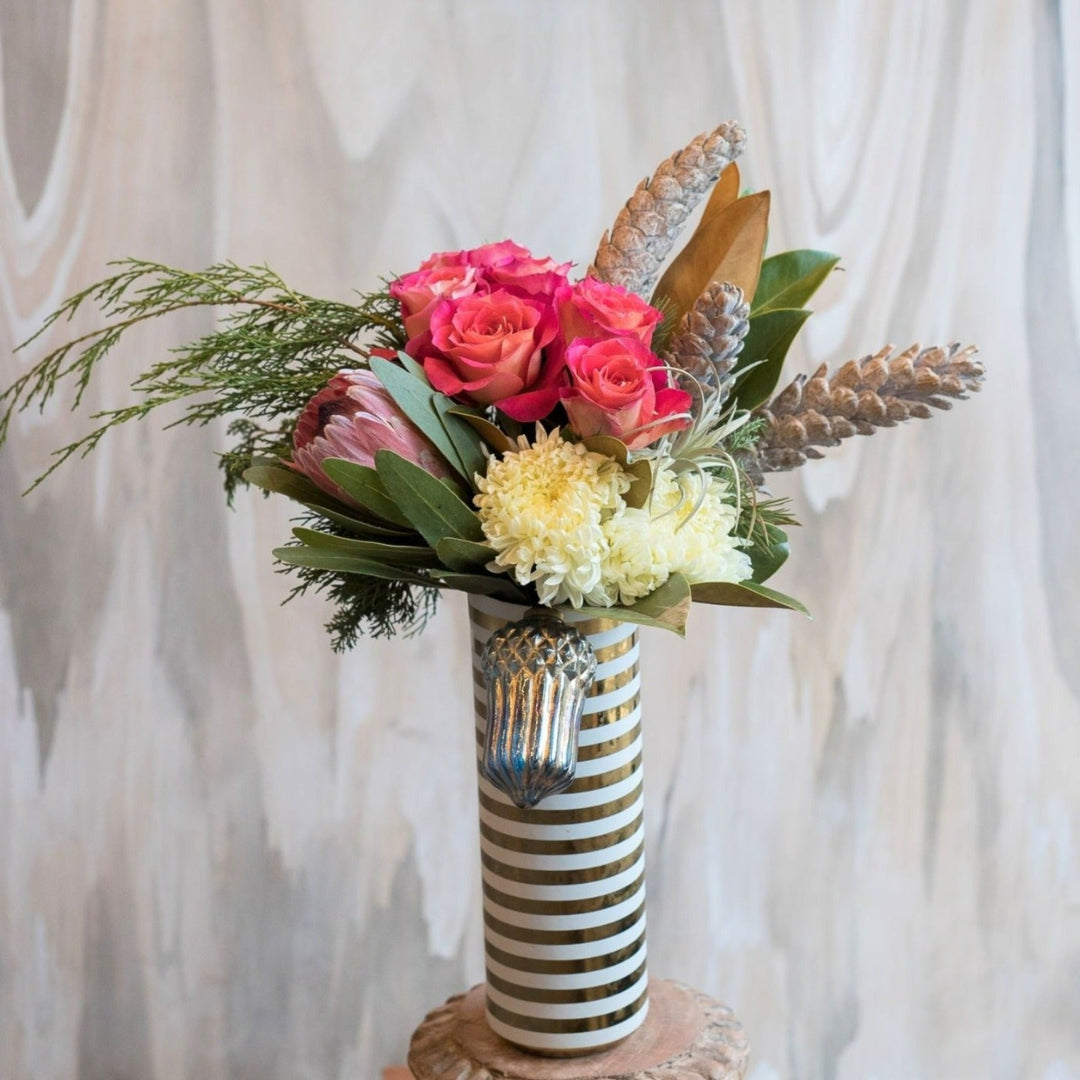 Woodland Wanderlust | A vased arrangement with a protea, roses, mums, evergreens, magnolia, pinecones, and a silver ornament in a striped vase.