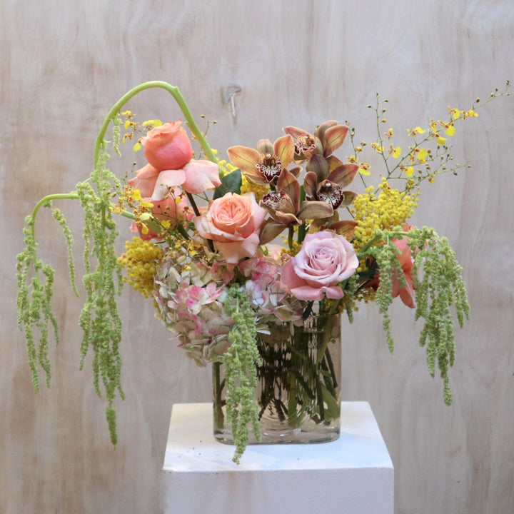 Sunbeam | An arrangement featuring roses, orchids, hydrangea, ameranthus, and other florals. Colors or gree, pink, and yellow. In a transparent vase. Photo taken against wooden background.