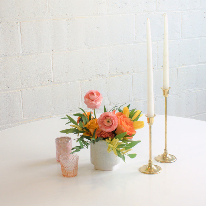 A floral centerpiece featuring a gold-colored candle, a white vase, and a selection of multicolored flowers including pink carnations and green foliage, placed on a white surface with a textured wall in the background.