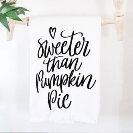 Festive Tea Towels | A white towel with black text that says "Sweeter the Pumpkin Pie". Towel is hanging on a wooden dowel with a plant in the background.