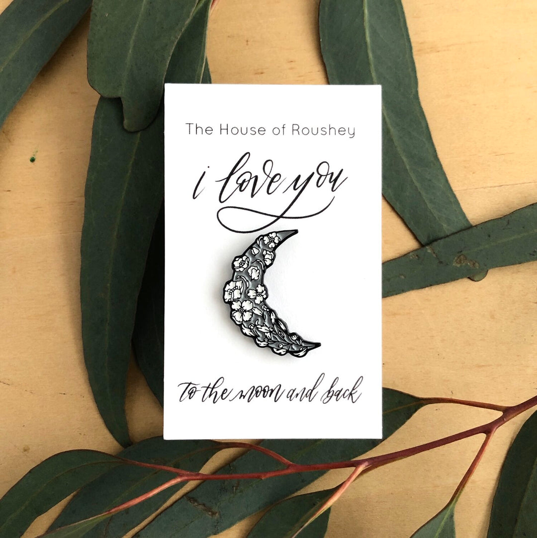 Enamel Pins by Christy Roushey | The House of Roushey, "I Love You To The Moon And Back". A gray enamel pin in the shape of a crescent moon decorated with white florals.