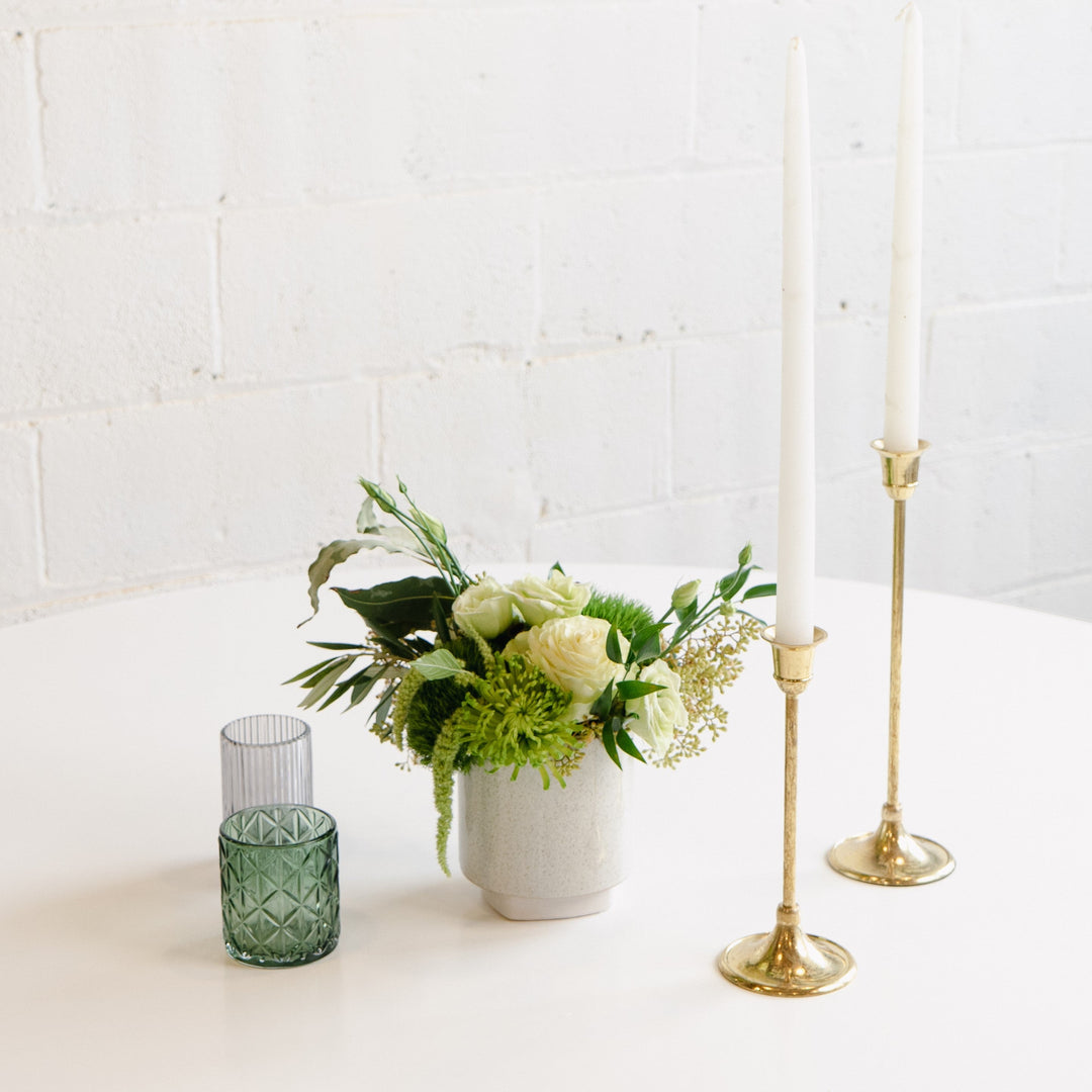 A floral centerpiece featuring a gold-colored candle, a white vase, and a selection of multicolored flowers including pink carnations and green foliage, placed on a white surface with a textured wall in the background.
