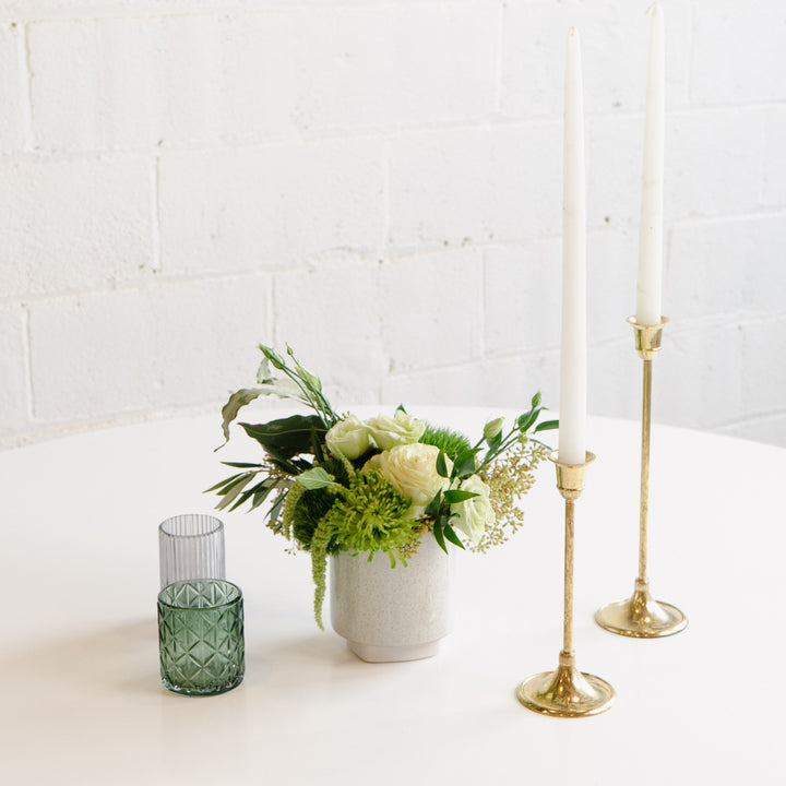 A floral centerpiece featuring a gold-colored candle, a white vase, and a selection of multicolored flowers including pink carnations and green foliage, placed on a white surface with a textured wall in the background.