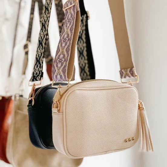 Willow Camera Crossbody Bag | Image shows the crossbody bags in black and cream. Focus on the cream bag. It has a pastel purple strap and gold accent hardware,