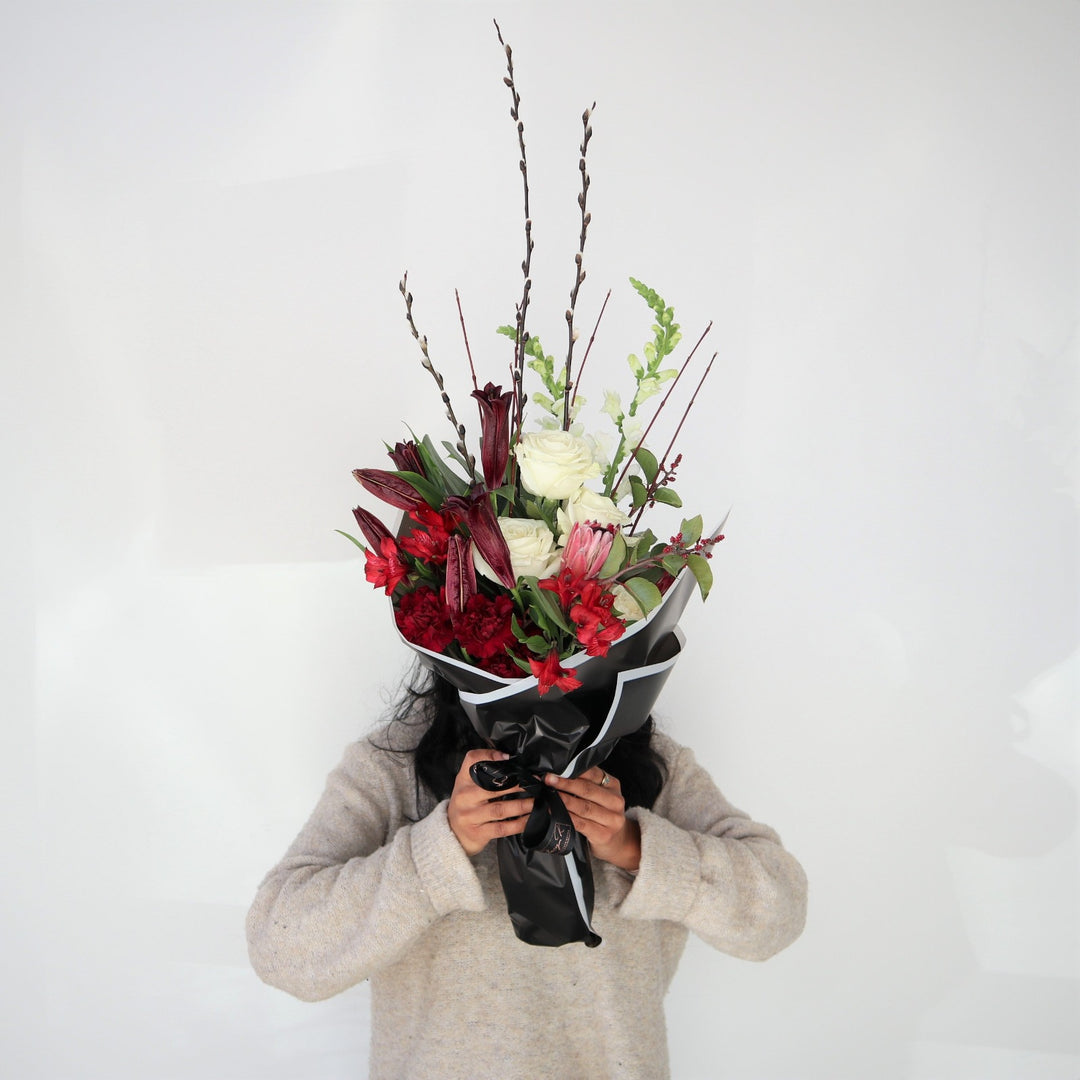 Red and White Wrap | Floral arrangement with white roses, and snap dragons, with red protea, carnations, other red flowers, and accent greens. Arrangement is being held up covering the model's face and is wrapped in black paper with a white edge.