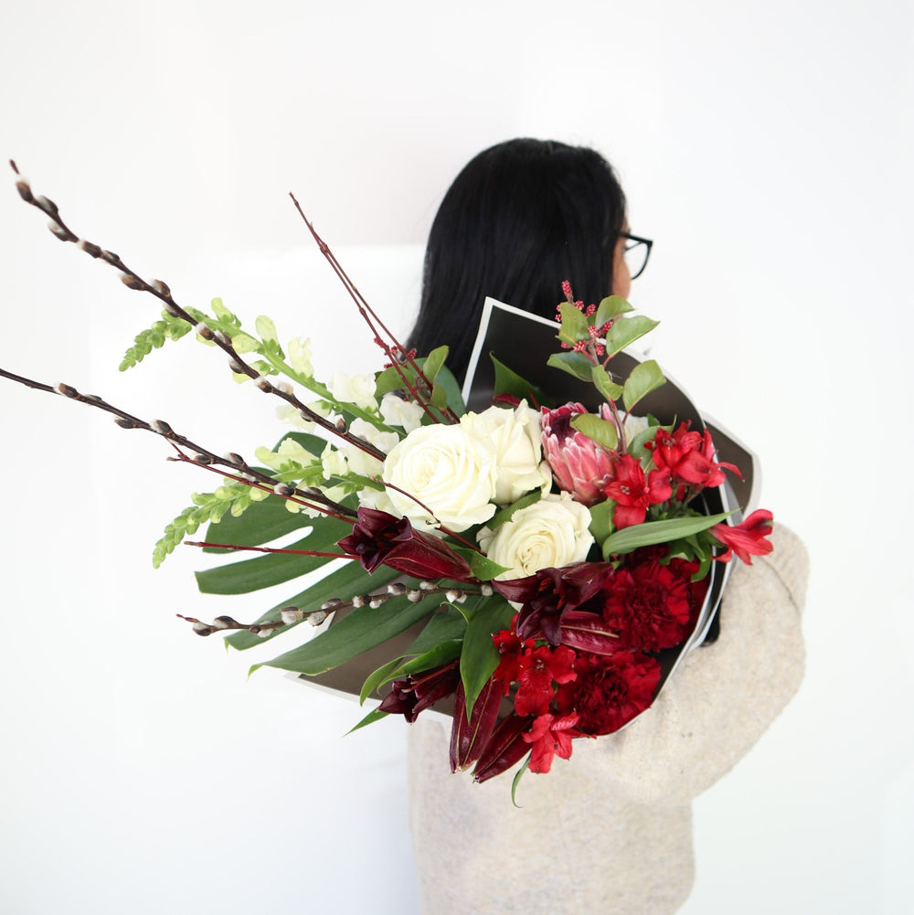 Red and White Wrap | Floral arrangement with white roses, and snap dragons, with red protea, carnations, other red flowers, and accent greens. Arrangement is being held over a model's shoulder, showing a different angle of the wrap and is wrapped in black paper with a white edge.