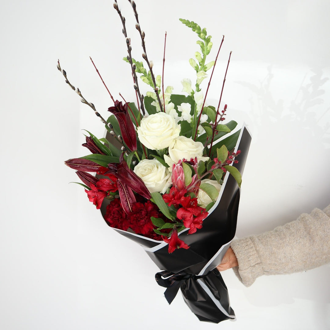 Red and White Wrap | Floral arrangement with white roses, and snap dragons, with red protea, carnations, other red flowers, and accent greens. Arrangement is being held against a backdrop and is wrapped in black paper with a white edge.
