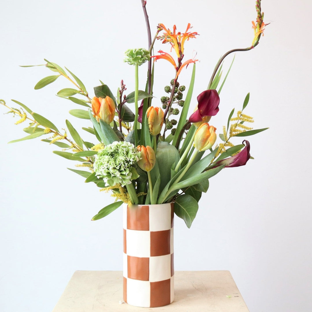 Adventure | A green and orange arrangement with hydrangea, tulips, and other florals. Arrangement is in a checkered orange and white vase. Close up on the arrangement.