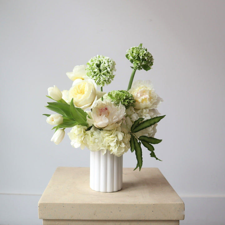 Limelight | A floral arrangement with whites, greens, and green blend florals. Arrangement is in a white vase on a cream pedestal against a white background.