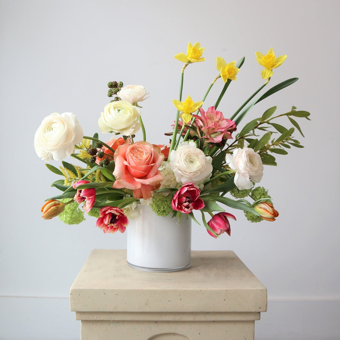 Day Dreaming | A colorful arrangement with whites, yellows, pinks, and oranges, with accents greens. Arrangement is in a white vase on a cream pedestal against a white background.