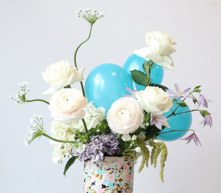 Happy | A white, green, purple, and blue arrangement with roses, accent balloons, and other florals. Vase is white with blue, gold, pink, and black specks. Up close photo of the arrangement.