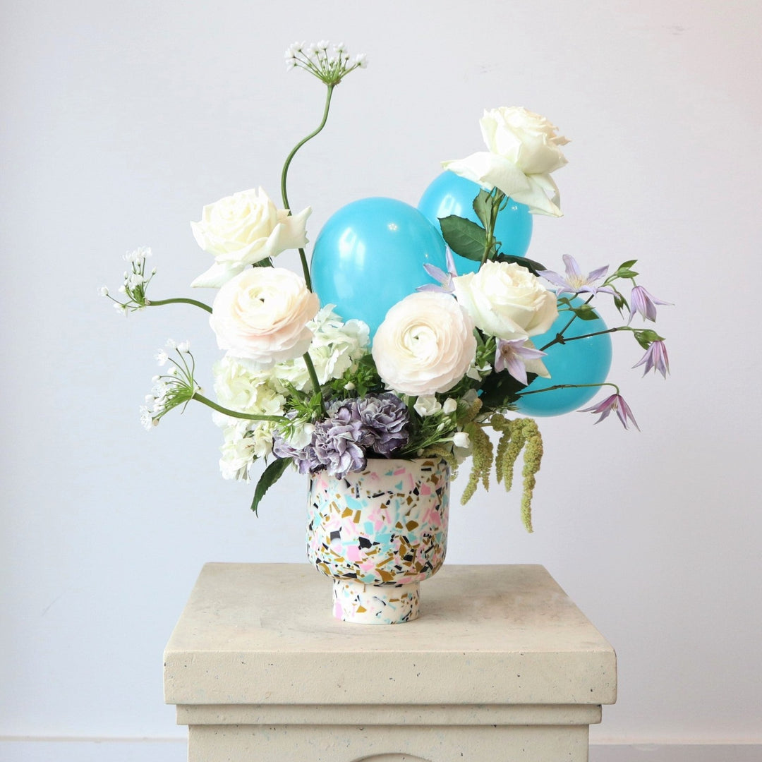 Happy | A white, green, purple, and blue arrangement with roses, accent balloons, and other florals. Vase is white with blue, gold, pink, and black specks. Photo taken on a pedestal against a white background.