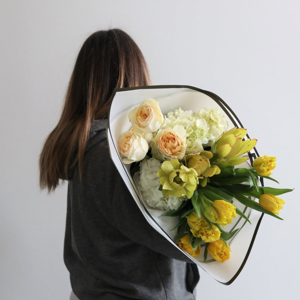 White paper around a floral arrangement, with flowers in it. The wrap has yellow tulips, yellow safari, peach roses and white hydrangea.
