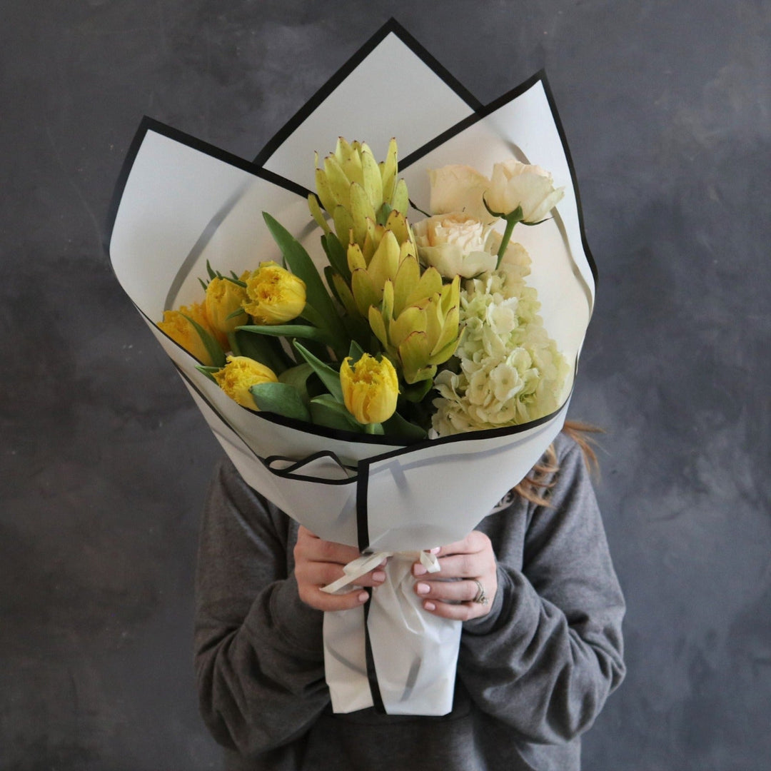 White paper around a floral arrangement, with flowers in it. The wrap has yellow tulips, yellow safari, peach roses and white hydrangea.