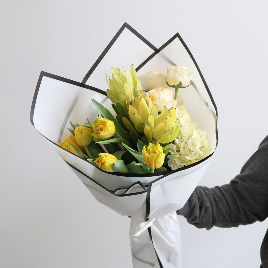 White paper around a floral arrangement, with flowers in it. The wrap has yellow tulips, yellow safari, peach roses and white hydrangea. 