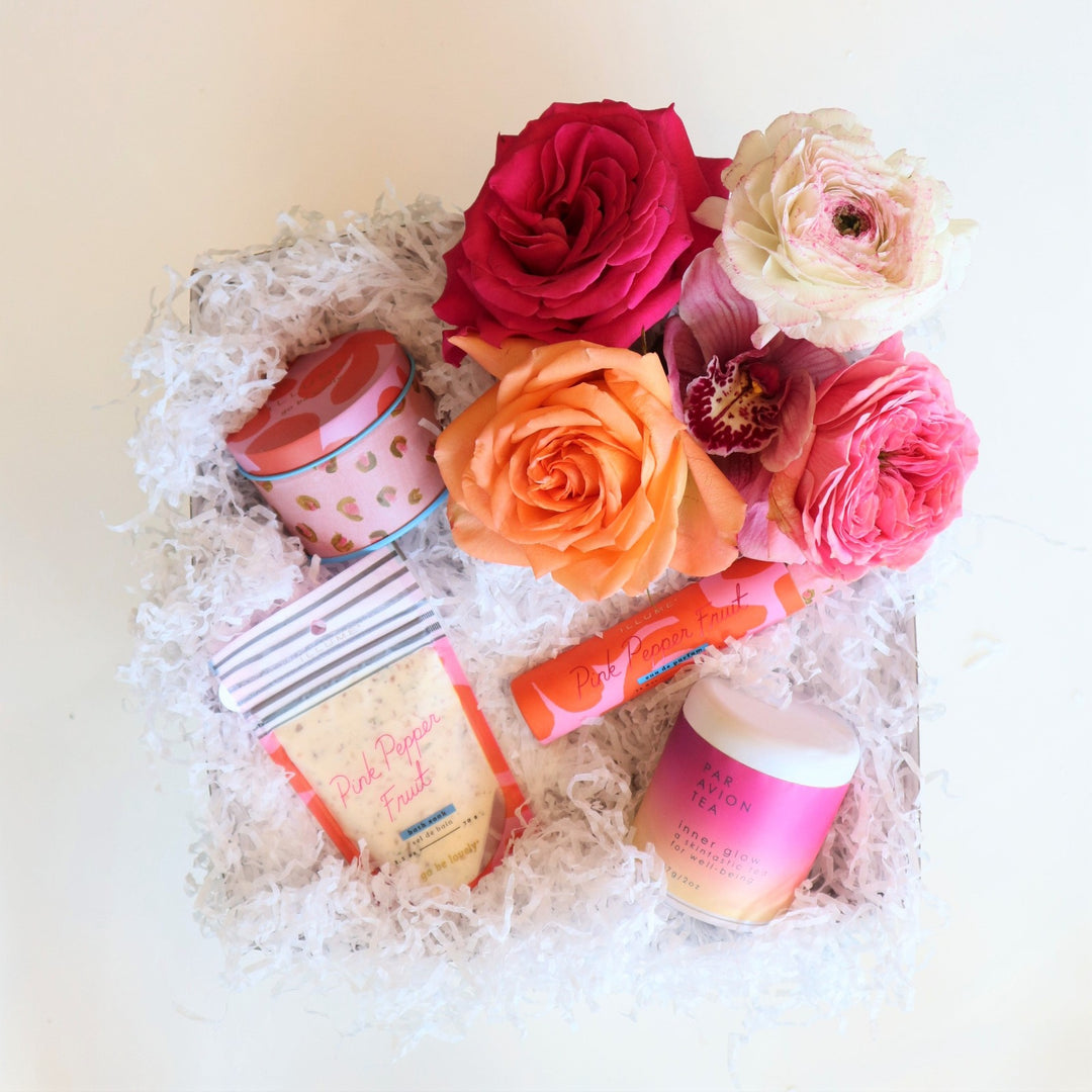 White shred in a square gift box with a bright rose and ranunculus vased arrangement. An ilume tin can candle with pink pattern on it. Pink Pepper fruit illume bath soak. Par Avion Tea in ombre pink and orange tin can. illume Pink Pepper Fruit roll on perfume.