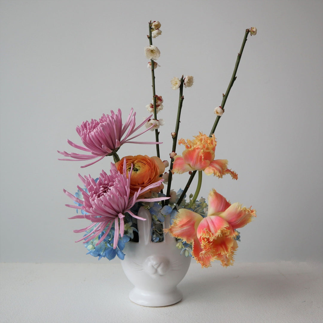 White bunny vase with a blue hydrangea, orange tulips, lavender mums, orange ranunculus and plum branching