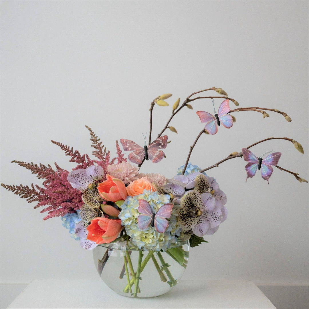 A vibrant floral arrangement featuring a variety of blooms and butterflies, displayed in a clear bowl.