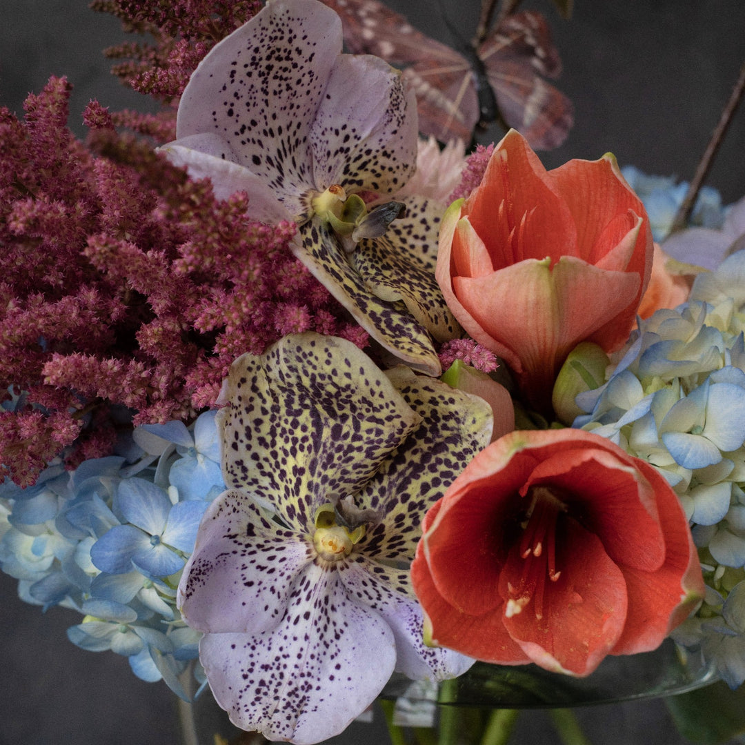 Whymsical floral arrangment with butterflies. Transparent bowl vase with Orchids, blue hydrangea, and decorative butterflies. Close up on Lavender speckled orchids.