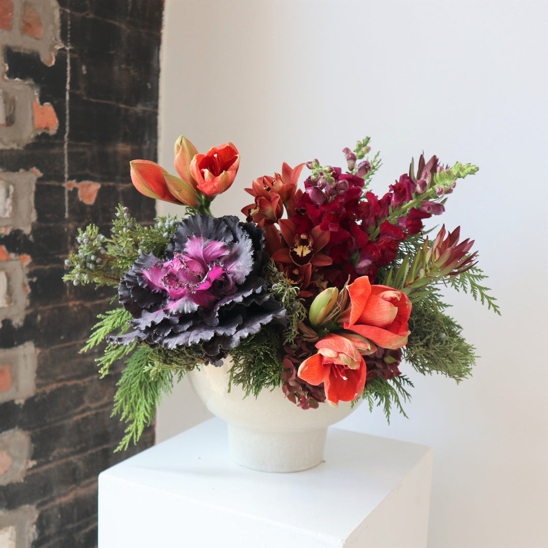 White bowl vase filled with flowers. Included in the arrangement are burgundy snapdragons, peach amaryllis, purple kale, orange cymbidium orchids and cedar evergreens.