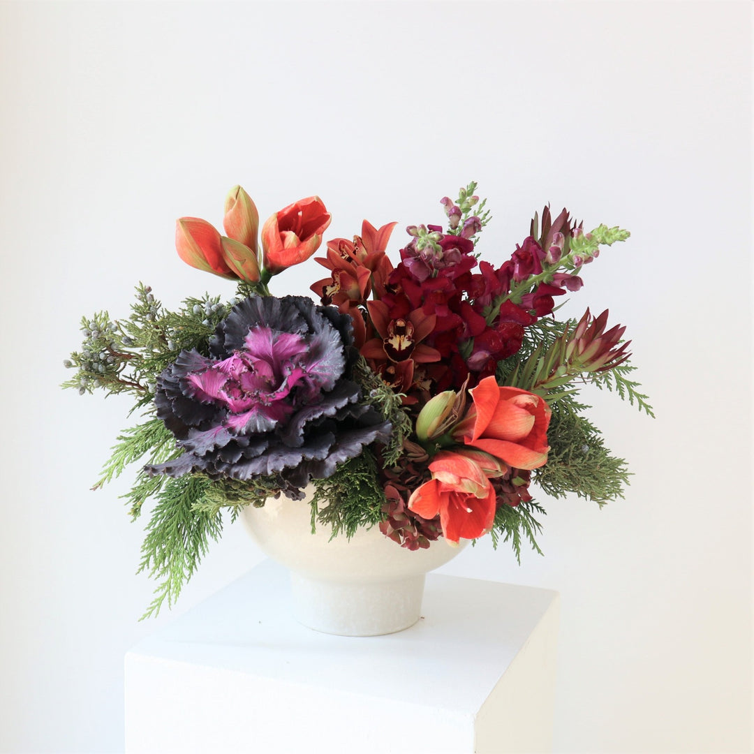 White bowl vase filled with flowers. Included in the arrangement are burgundy snapdragons, peach amaryllis, purple kale, orange cymbidium orchids and cedar evergreens.
