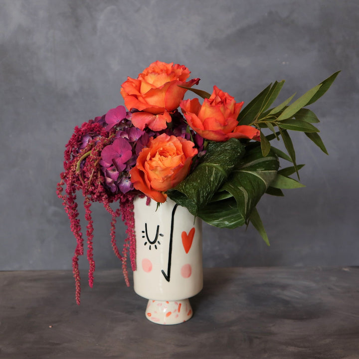 Face vase with blushing cheek a winking eye and one heart eye, with three orange roses, one purple hydrangea, greenery and red hanging amaranthus. 