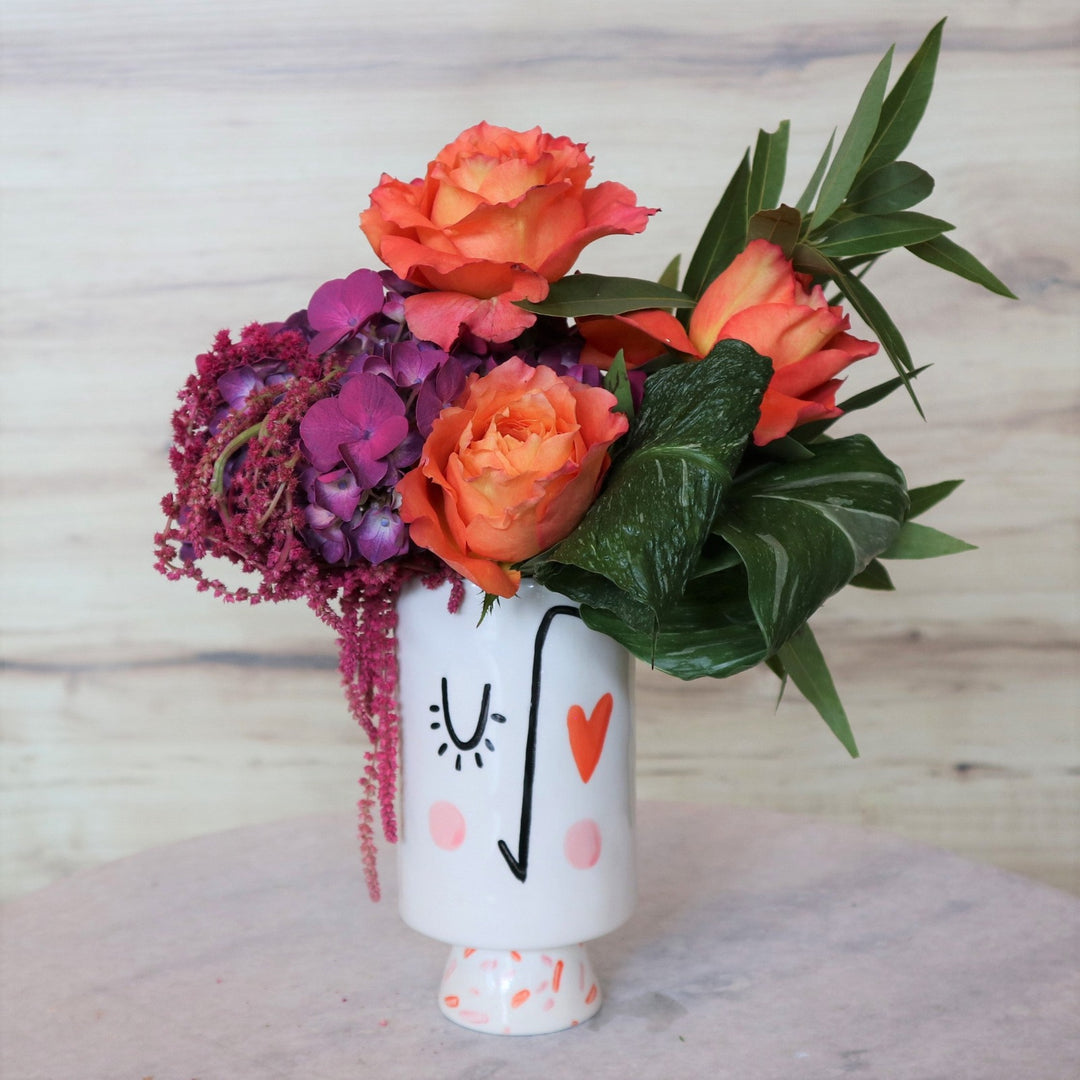 Face vase with blushing cheek a winking eye and one heart eye, with three orange roses, one purple hydrangea, greenery and red hanging amaranthus.