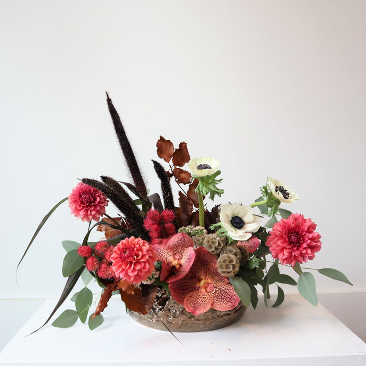 Arrangement in low bronze vase, pink dahlias, pink orchids, Red castor bean pods, red and green leaves, black millet, scaviosa pods, and white anemone flowers. Photo taken on white background.