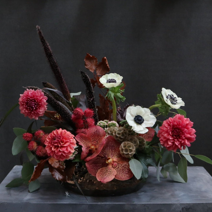 Arrangement in low bronze vase, pink dahlias, pink orchids, Red castor bean pods, red and green leaves, black millet, scaviosa pods, and white anemone flowers. Photo taken on gray pedestal with black background.