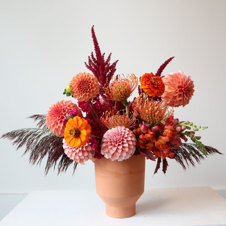 Arrangement in terracotta pot with pink and peach dahlias, pincushion protea, Pink ameranthus, orange/pink snapdragons, and purple grass. Photo taken with white background.