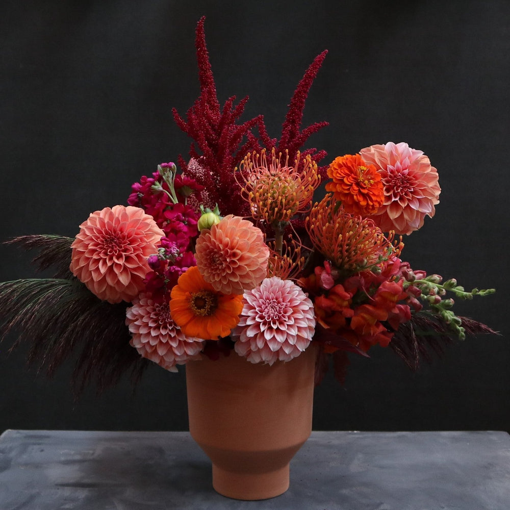 Arrangement in terracotta pot with pink and peach dahlias, pincushion protea, Pink ameranthus, orange/pink snapdragons, and purple grass. Photo taken on gray pedestal with Black background.