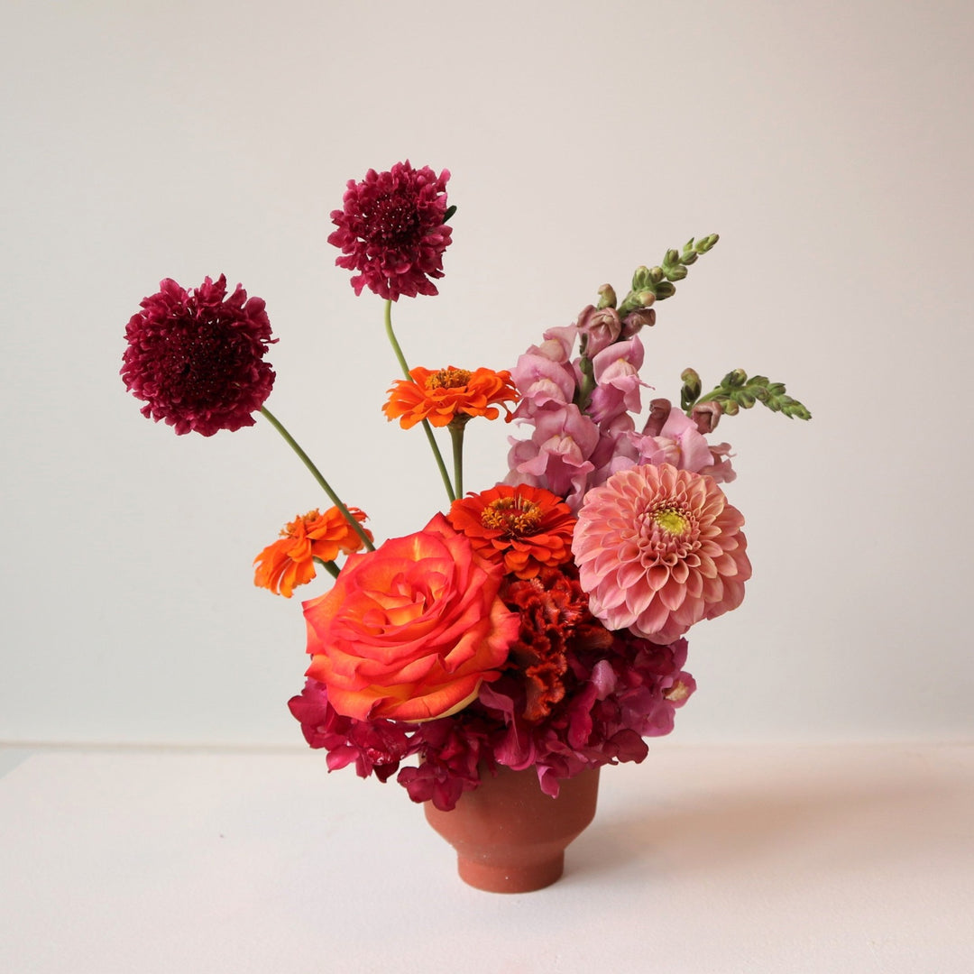 Flower arrangement with one orange rose, three orange zinnias, one pink dahlia, two pink snapdragons, pink hydrangea and two scabiosa in a small terracotta pot. 