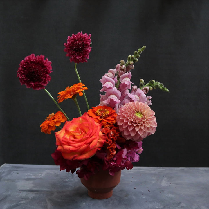 Flower arrangement with one orange rose, three orange zinnias, one pink dahlia, two pink snapdragons, pink hydrangea and two scabiosa in a small terracotta pot.
