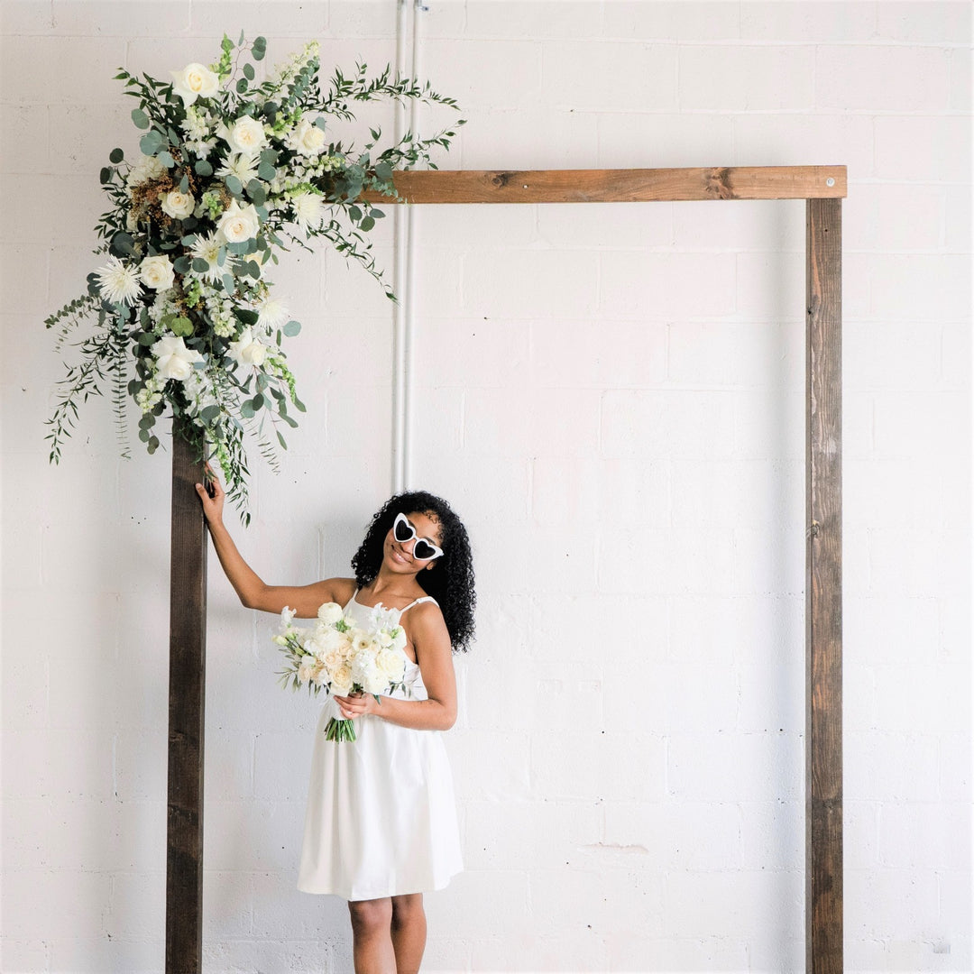 Flowered Arch Piece | A white and green arch arrangement with white roses, snapdragons, eucalyptus, and other greens/white florals. Arch piece is on a natural wood arch. Model is wearing a white dress, heart sunglasses, and holding a white bridal bouquet. 