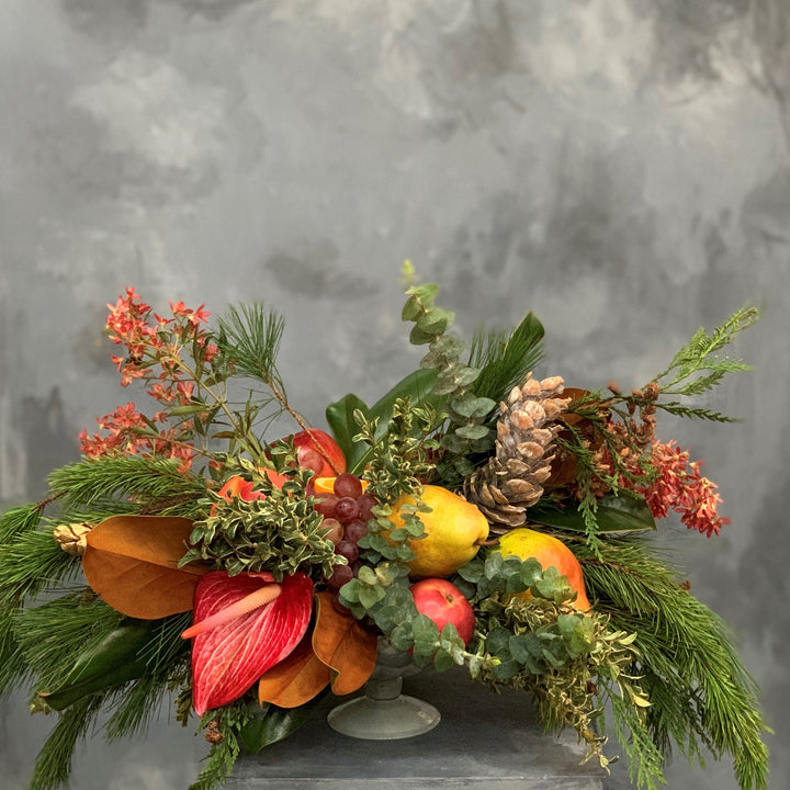 Fruited Evergreen Centerpiece. Grapes, mango, pears, and apples, with greens,  florals, and pinecones. Photo taken on gray background.