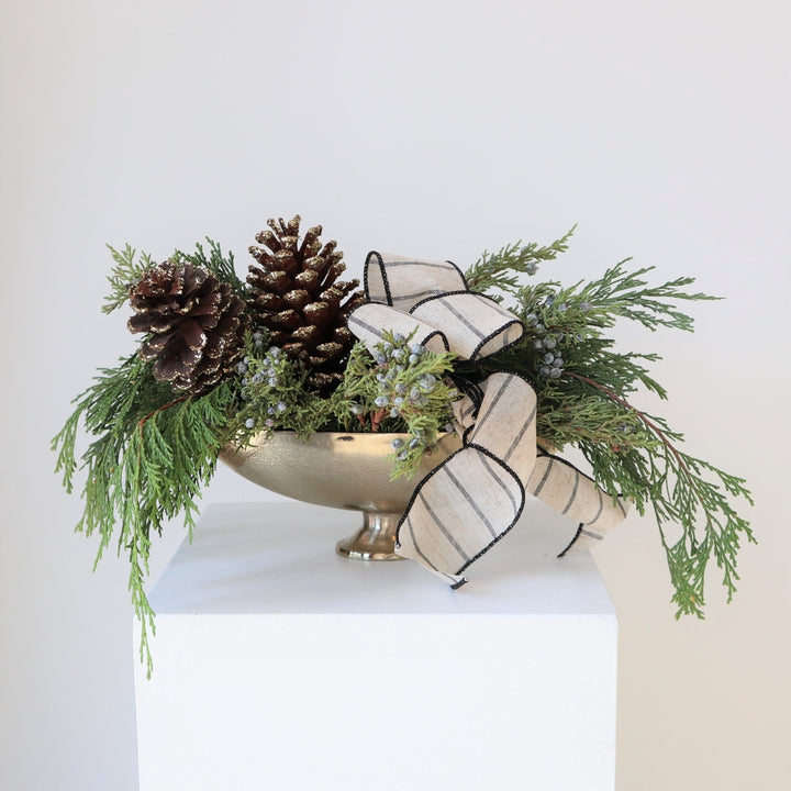 Arrangement in gold boat vase, with evergreens, glitter brown and gold pinecones and cream and black striped bow taken on a white background.