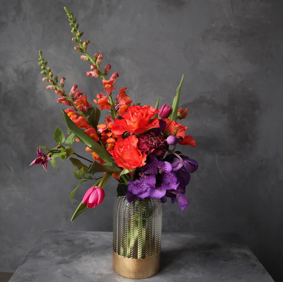 Gray background with orange roses, orange snapdragons, purple vanda orchids, pink tulips designed in a clear vase with a gold rim bottom.