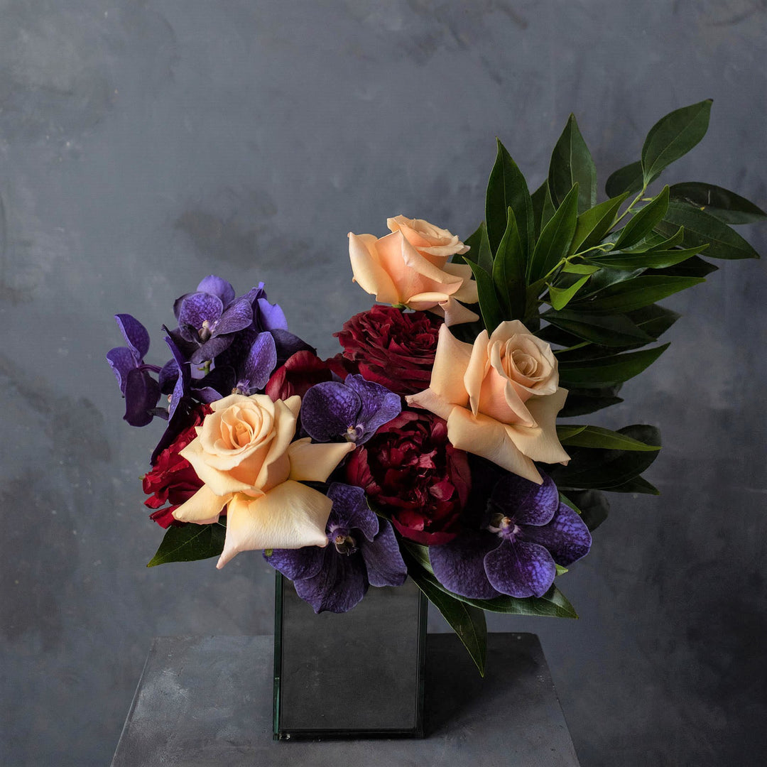 Mirrored vase with a floral piece in peach, red and purple accented with rich greens.  Rochester Contemporary Florist