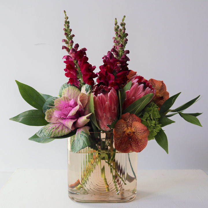 Taupe vase filled with kale, protea, snapdragons and orchids.