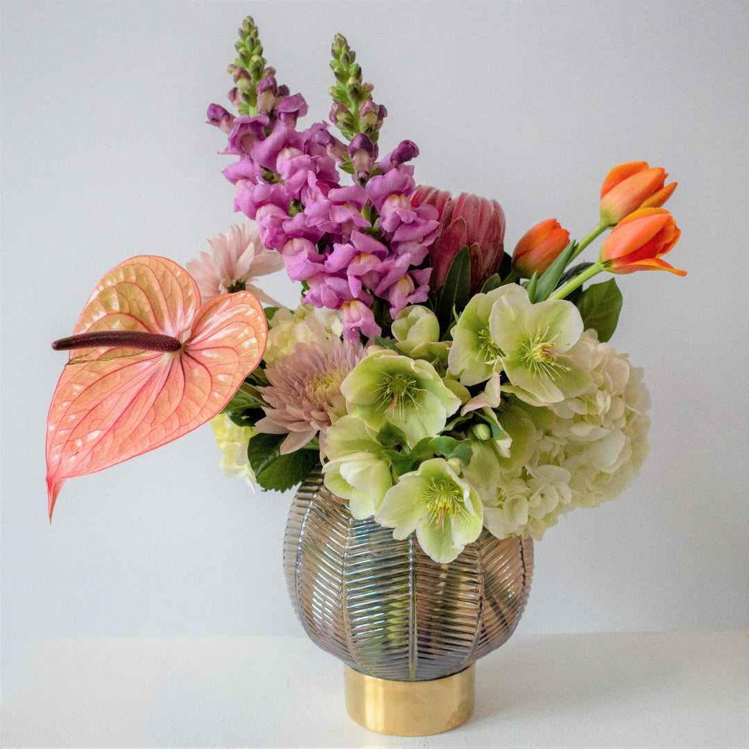 White background with vased arrangement that has green helleborus, lilac colored mums, purple snapdragons, orange tulips and a peach colored anthurium.