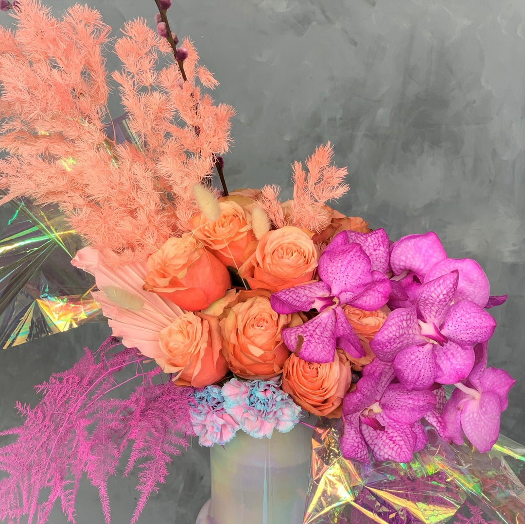 Whymsical Arrangement with orange roses, purple/pink orchids, colored ferns, and iridescent cellophane in and iridescent vase.