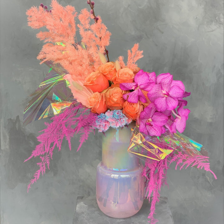 Whymsical Arrangement with orange roses, purple/pink orchids, colored ferns, and iridescent cellophane in and iridescent vase.