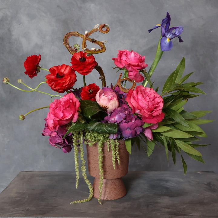 Dark background with Floral arrangement in front. Deep pink vase filled with pink roses, red ranunculus, one iris, purple hydrangea, green amaranthus, greenery, pussy willow and one pink peony.