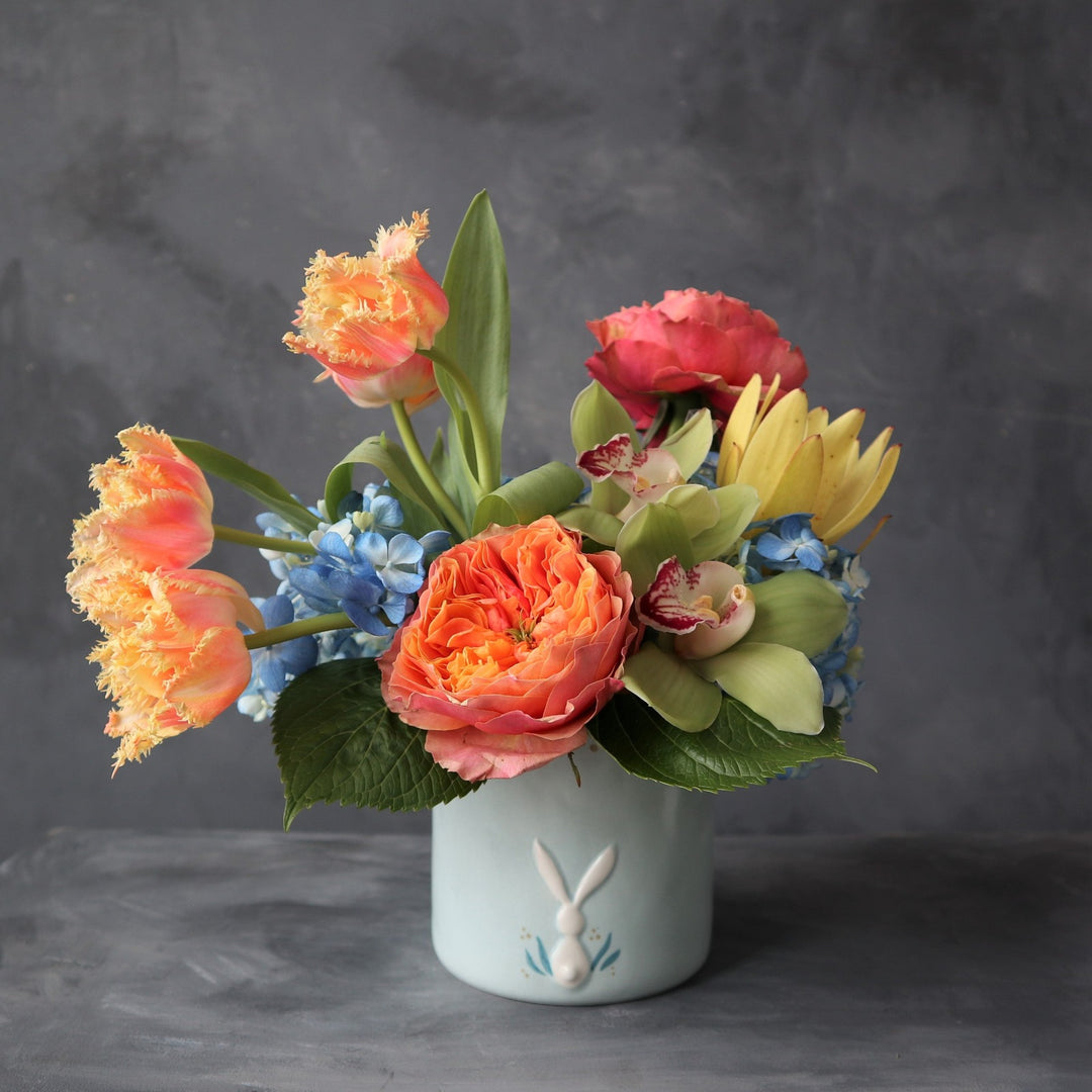 Orange garden roses, orange tulips, blue hydrangea, green orchids, yellow safari protea in a blue pot with white bunny on it. Photo taken on dark background.