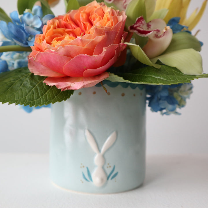 Orange garden roses, orange tulips, blue hydrangea, green orchids, yellow safari protea in a blue bunny pot.