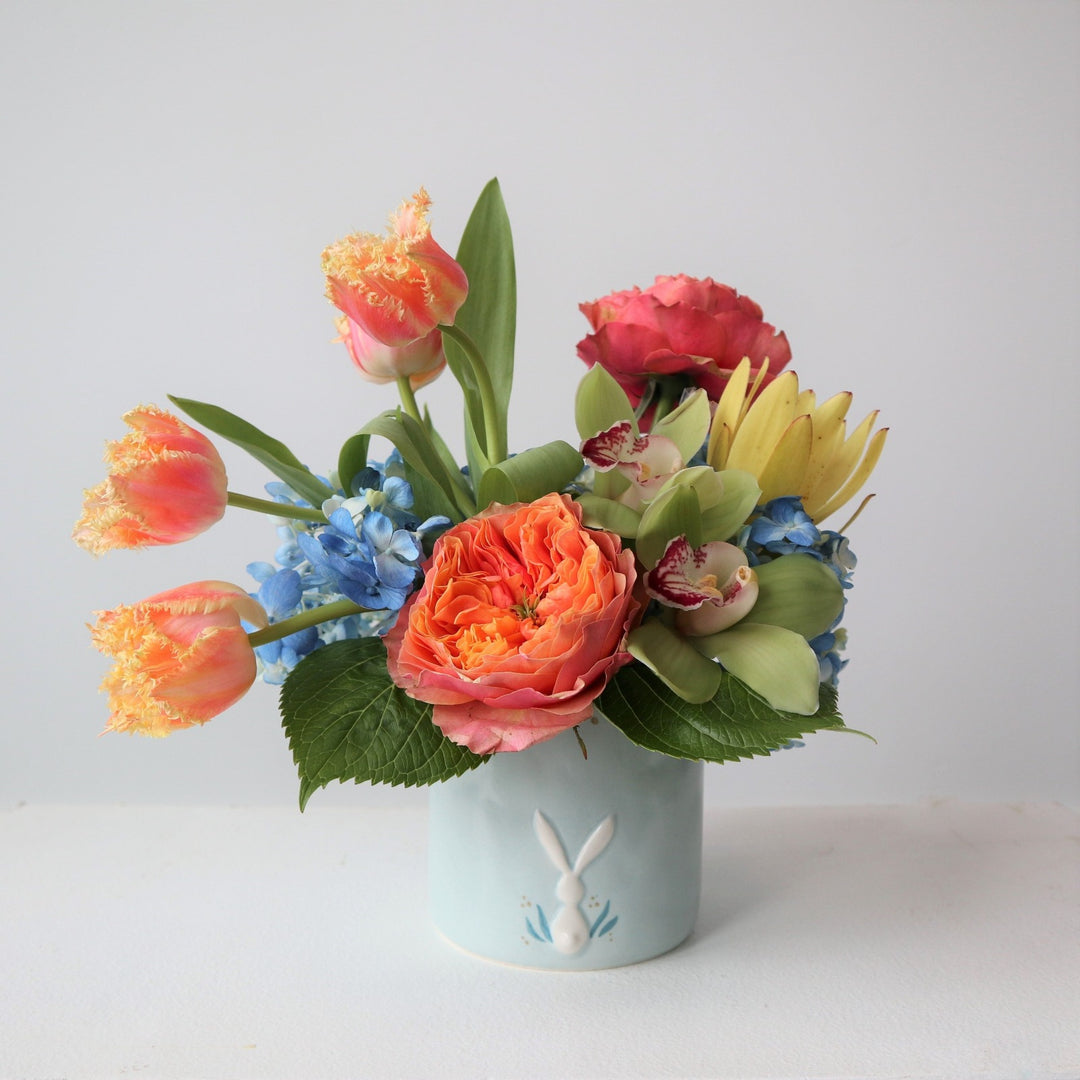 Orange garden roses, orange tulips, blue hydrangea, green orchids, yellow safari protea in a blue pot with white bunny on it. Photo taken on white background.