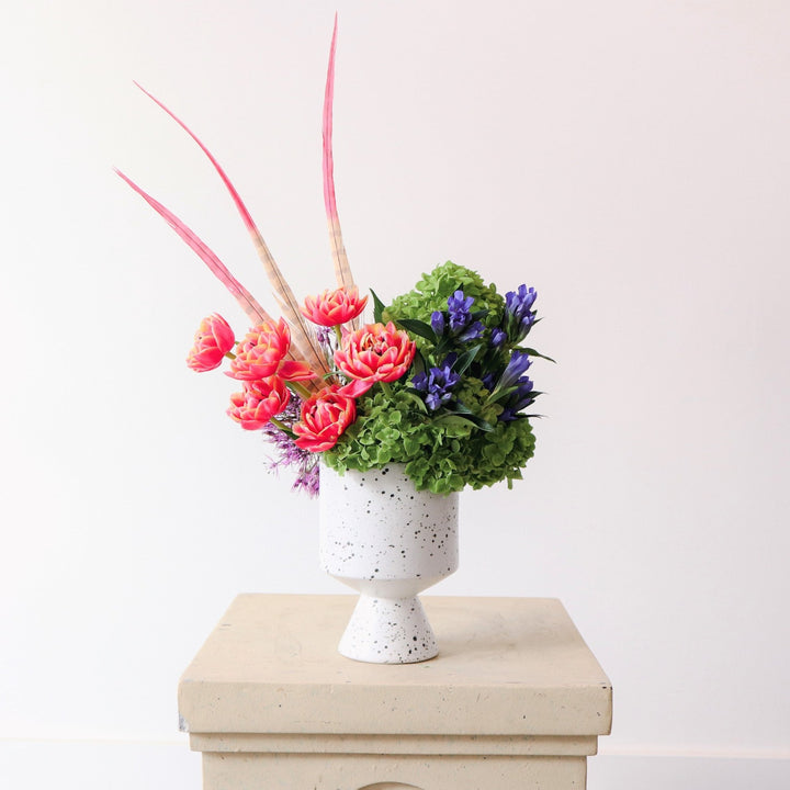 Filled with colorful Bloom | An arrangement filled with tulips, purple allium and green hydrangea. Accent flowers add asymmetry to the piece. Made in a white vase with black speckles. 