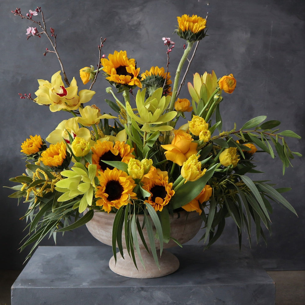 Yellow sunflowers, protea, orchids, roses and greenery.  Vased in a cream compote. 