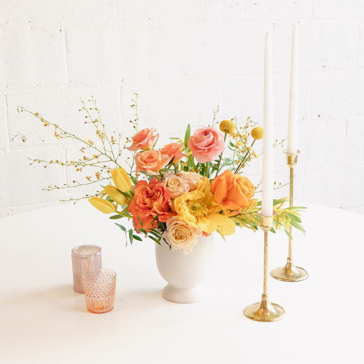 A floral centerpiece arrangement featuring a white vase, gold-colored candle holders, and a selection of purple, pink, and white flowers, placed on a white surface with a plain background.