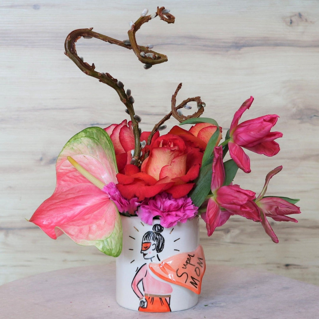 Light background with arrangement that is a girl with a mask and bun with a cape that says "super MOM". Flowers in the vase are pink tulips, coral roses, onepink anthurium and two pussy willow branches